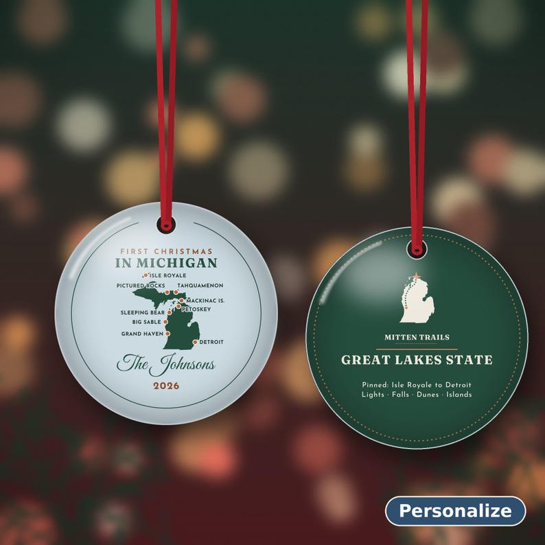 May include: Two round ceramic ornaments with red hanging strings. The left ornament is light blue with a map of Michigan, text reading First Christmas in Michigan, The Johnsons, and 2026. The right ornament is dark green with a white map outline.