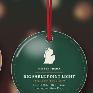 May include: A round, dark green ornament hanging from a red ribbon. It features a white map of Michigan with a star, the text "MITTEN TRAILS", "BIG SABLE POINT LIGHT", coordinates, "First lit 1867", "112 ft tower", and "Ludington State Park".