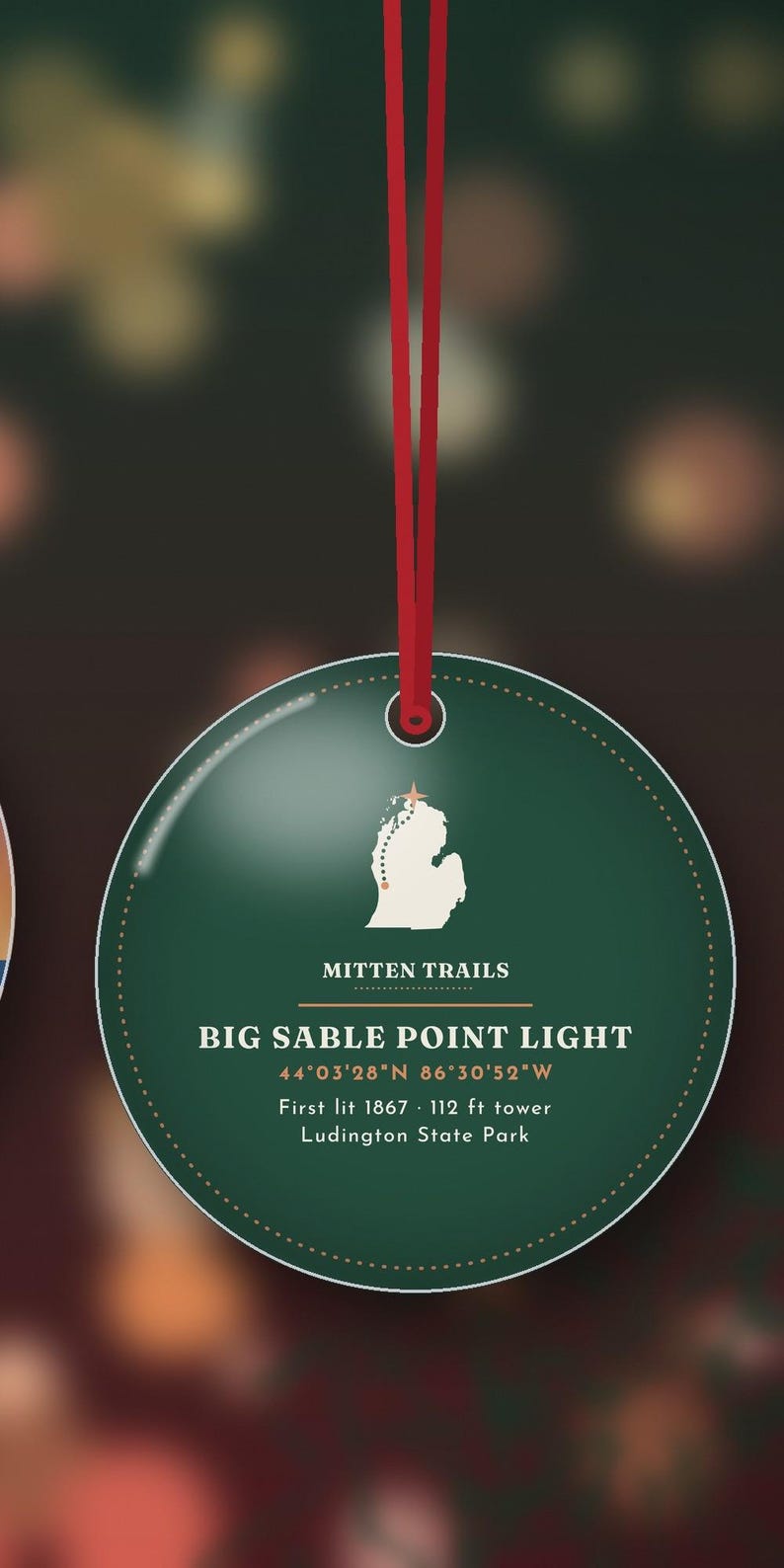 May include: A round, dark green ornament hanging from a red ribbon. It features a white map of Michigan with a star, the text "MITTEN TRAILS", "BIG SABLE POINT LIGHT", coordinates, "First lit 1867", "112 ft tower", and "Ludington State Park".