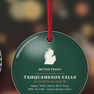 May include: A round, dark green ornament hanging from a red ribbon. It features a white map of Michigan with a star, the text "MITTEN TRAILS", "TAHQUAMENON FALLS", coordinates, and details: "Upper Falls: about 48 ft drop, 200+ ft wide, tannin-brown water."
