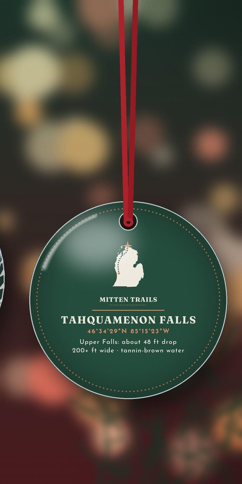 May include: A round, dark green ornament hanging from a red ribbon. It features a white map of Michigan with a star, the text "MITTEN TRAILS", "TAHQUAMENON FALLS", coordinates, and details: "Upper Falls: about 48 ft drop, 200+ ft wide, tannin-brown water."