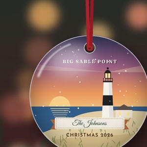 May include: A round, flat ornament hanging from a red ribbon, featuring a graphic of the Big Sable Point lighthouse at sunset. The design includes the text BIG SABLE POINT, The Johnsons, and CHRISTMAS 2026. The scene depicts a beach, ocean, and sky.