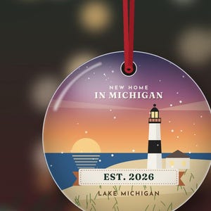 May include: A round ceramic ornament hanging from a red ribbon, featuring a sunset illustration of a lighthouse on a beach. The text reads NEW HOME IN MICHIGAN, EST. 2026, and LAKE MICHIGAN. The design includes a blue sky, orange horizon, and sand.