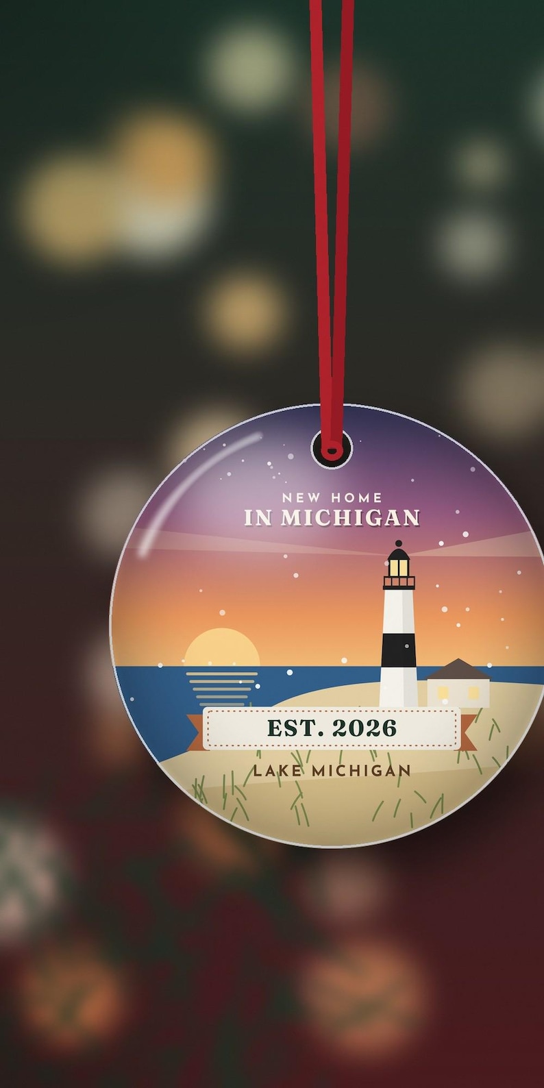 May include: A round ceramic ornament hanging from a red ribbon, featuring a sunset illustration of a lighthouse on a beach. The text reads NEW HOME IN MICHIGAN, EST. 2026, and LAKE MICHIGAN. The design includes a blue sky, orange horizon, and sand.