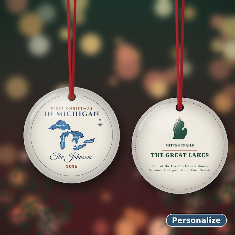 May include: Two round white ceramic ornaments with red ribbon hangers. The left ornament features a blue map of the Great Lakes, the text First Christmas In Michigan, The Johnsons, and 2026. The right ornament shows a green Michigan map, Mitten Trails, and The Great Lakes text.