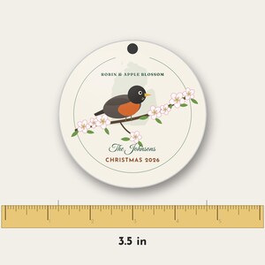 May include: A round, cream-colored ornament featuring a robin perched on an apple blossom branch. Text reads ROBIN & APPLE BLOSSOM, The Johnsons, and CHRISTMAS 2026. A ruler below shows the ornament has a diameter of 3.5 inches.