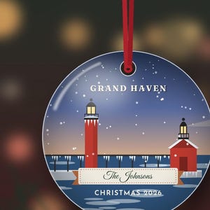 May include: A round, flat Christmas ornament hanging from a red ribbon. The design features a winter scene with the Grand Haven lighthouse, a pier, and snow. It includes the text 'GRAND HAVEN', 'The Johnsons', and 'CHRISTMAS 2026'.