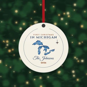 May include: A round, cream-colored ceramic ornament hanging from a red ribbon. It features a blue map of the Great Lakes, the text 'FIRST CHRISTMAS IN MICHIGAN', 'The Johnsons', the year '2026', and a small compass rose icon.