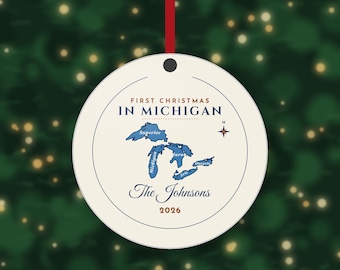 May include: A round, cream-colored ceramic ornament hanging from a red ribbon. It features a blue map of the Great Lakes, the text 'FIRST CHRISTMAS IN MICHIGAN', 'The Johnsons', the year '2026', and a small compass rose icon.