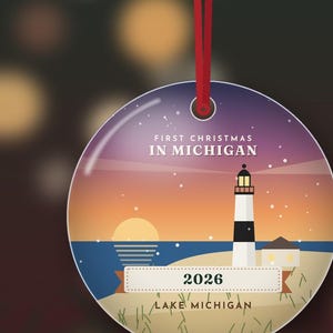 Op de afbeelding: Een ronde keramische hanger aan een rood lint, met een illustratie van een zonsondergang met een vuurtoren op een strand. De tekst luidt FIRST CHRISTMAS IN MICHIGAN, 2026 en LAKE MICHIGAN. Het ontwerp bevat een zwart-witte vuurtoren.