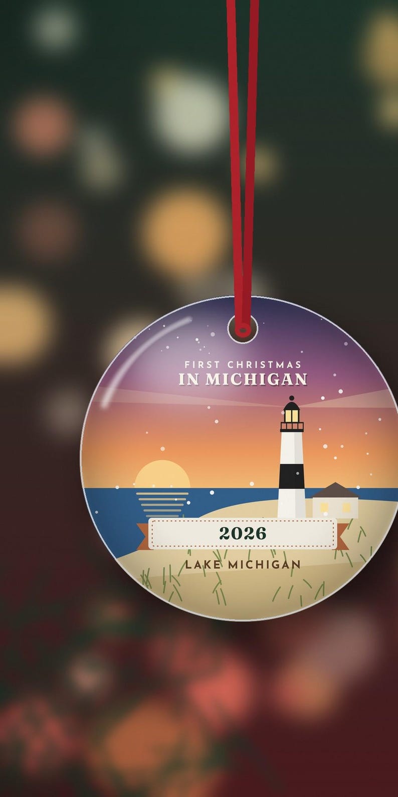 Op de afbeelding: Een ronde keramische hanger aan een rood lint, met een illustratie van een zonsondergang met een vuurtoren op een strand. De tekst luidt FIRST CHRISTMAS IN MICHIGAN, 2026 en LAKE MICHIGAN. Het ontwerp bevat een zwart-witte vuurtoren.