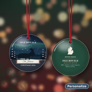 May include: Two round, flat Christmas ornaments with red hanging ribbons. The left ornament features a dark blue night scene with a moose, pine trees, and the text "ISLE ROYALE", "The Johnsons", and "CHRISTMAS 2026". The right ornament is green with a white map outline, "MITTEN TRAILS", "ISLE ROYALE", coordinates, and text about Lake Superior.