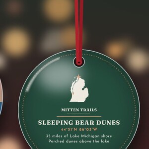 Puede incluir: Un adorno colgante redondo de color verde oscuro con una cinta roja. Presenta un mapa blanco de Michigan con una estrella, el texto "MITTEN TRAILS", "SLEEPING BEAR DUNES", "44&deg;51'N 86&deg;03'W", "35 miles of Lake Michigan shore" y "Perched dunes above the lake."