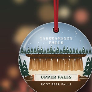 Puede incluir: Un adorno redondo de cer&aacute;mica colgado de una cinta roja, con una ilustraci&oacute;n gr&aacute;fica de Tahquamenon Falls. El dise&ntilde;o incluye una cascada marr&oacute;n, pinos verdes y el texto Tahquamenon Falls, Upper Falls y Root Beer Falls.