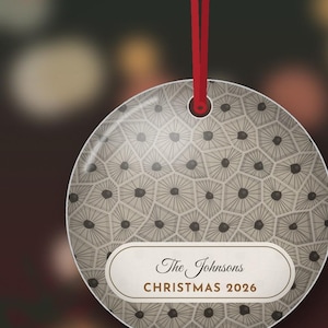 May include: A round, flat, beige-toned ornament featuring a geometric pattern of dark-centered hexagons. A white horizontal label in the center displays the text "The Johnsons" and "CHRISTMAS 2026". A thin red ribbon is attached to the top.