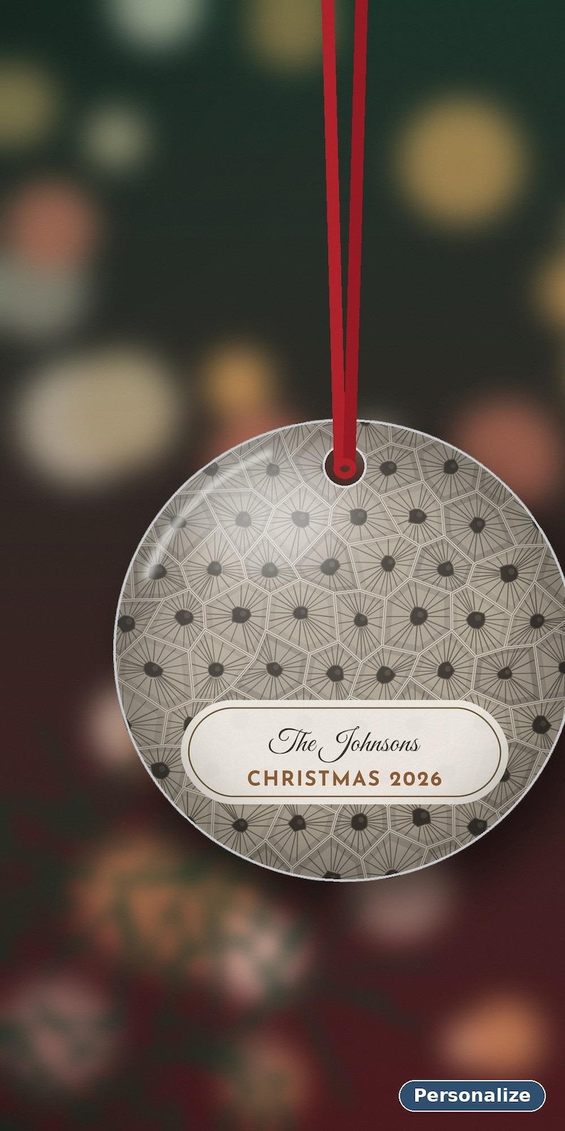 May include: A round, flat, beige-toned ornament featuring a geometric pattern of dark-centered hexagons. A white horizontal label in the center displays the text "The Johnsons" and "CHRISTMAS 2026". A thin red ribbon is attached to the top.
