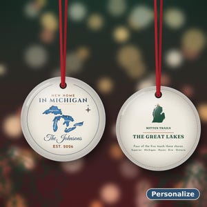 May include: Two round, cream-colored ceramic ornaments with red hanging ribbons. The left ornament features a blue map of the Great Lakes, text reading New Home In Michigan, The Johnsons, and Est. 2026. The right shows a green Michigan map, Mitten Trails, and The Great Lakes text.
