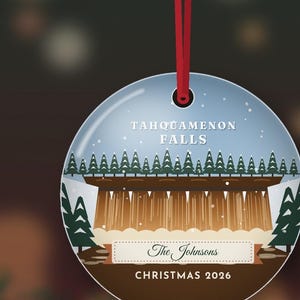 May include: A round, flat, white ceramic ornament hanging from a red ribbon. It features a graphic of Tahquamenon Falls with green pine trees, brown water, and white snow. Text reads: TAHQUAMENON FALLS, The Johnsons, and CHRISTMAS 2026.