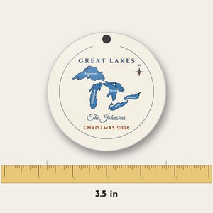 May include: A round white ornament featuring a blue map of the Great Lakes with labeled names, a compass rose, and the text "The Johnsons" and "CHRISTMAS 2026". A ruler below shows the ornament has a diameter of 8.89 cm.