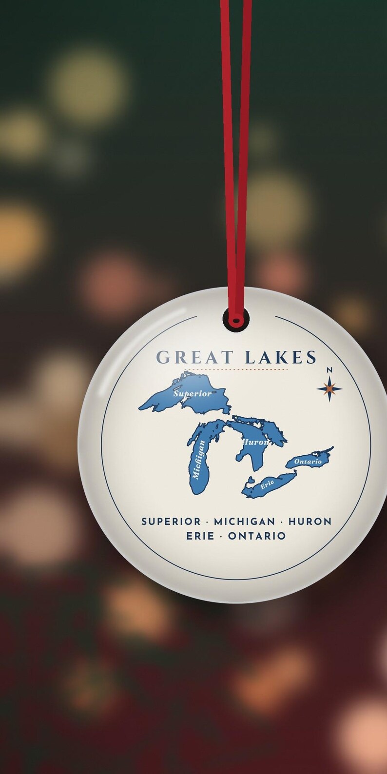 May include: A round white ceramic ornament hanging from a red ribbon. The center features a blue map of the Great Lakes labeled Superior, Michigan, Huron, Erie, and Ontario. The text GREAT LAKES is printed above, with the names listed below.