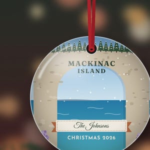 May include: A round, flat Christmas ornament hanging from a red ribbon. The design features a stylized illustration of Mackinac Island with green pine trees, a blue sky, and water. It includes the text "MACKINAC ISLAND", "The Johnsons", and "CHRISTMAS 2026".