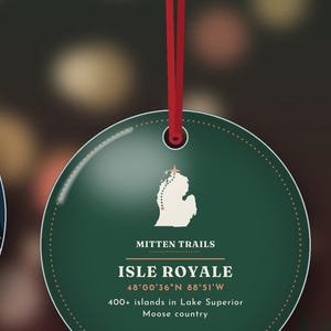 May include: A round, dark green ornament hanging from a red ribbon. The design features a white map of Isle Royale with a small star, the text MITTEN TRAILS, ISLE ROYALE, 48&deg;00'36"N 88&deg;51'W, and 400+ islands in Lake Superior Moose country.