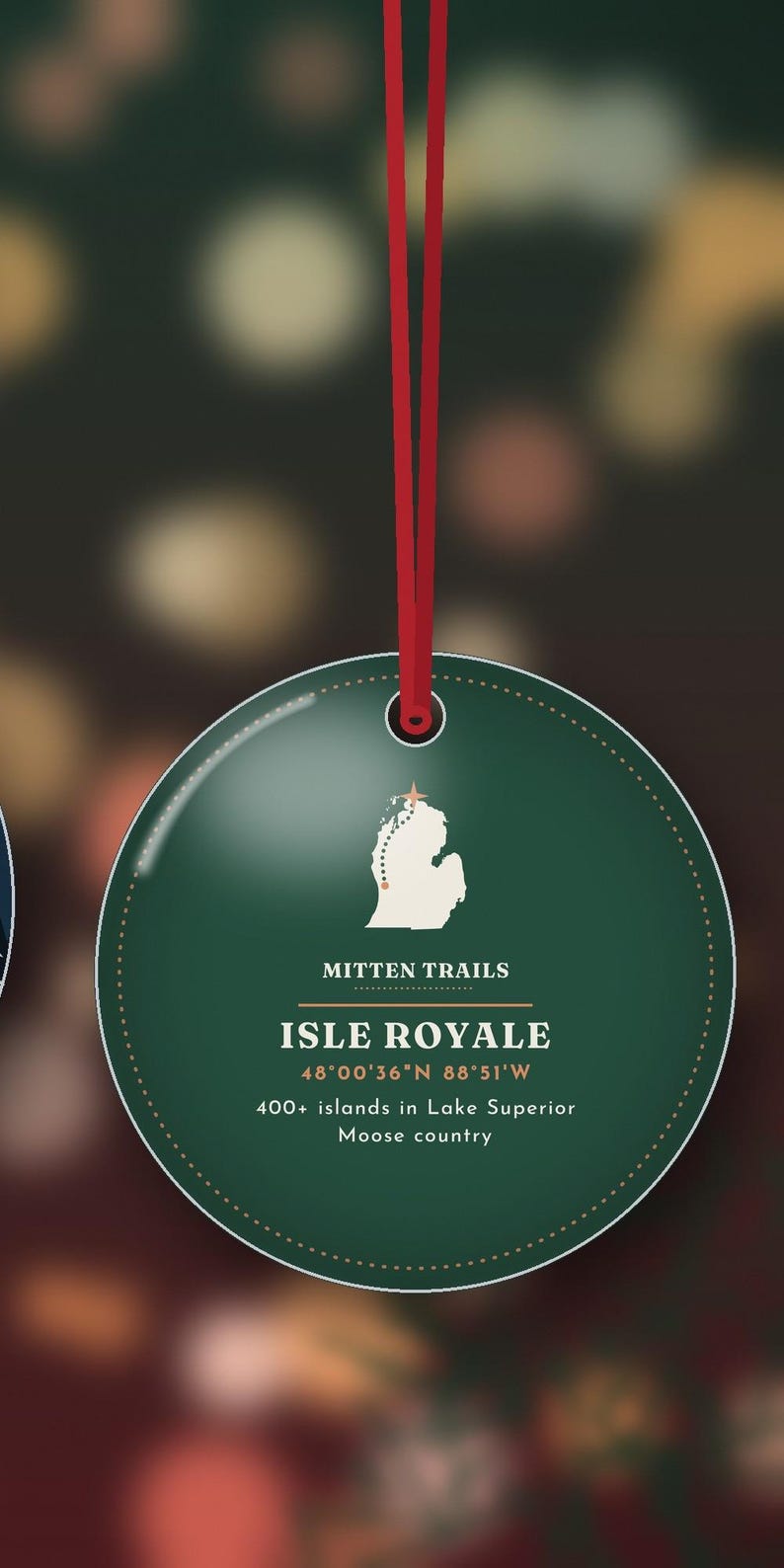 May include: A round, dark green ornament hanging from a red ribbon. The design features a white map of Isle Royale with a small star, the text MITTEN TRAILS, ISLE ROYALE, 48&deg;00'36"N 88&deg;51'W, and 400+ islands in Lake Superior Moose country.