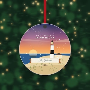 May include: A round Christmas ornament featuring a lighthouse on a beach at sunset with a purple starry sky. The text reads FIRST CHRISTMAS IN MICHIGAN, The Johnsons, and 2026. The ornament hangs from a red ribbon against a blurred green background.