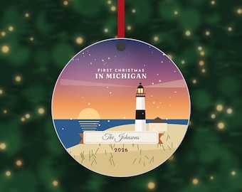 Puede incluir: Un adorno navideño redondo con un faro en una playa al atardecer bajo un cielo estrellado púrpura. El texto dice FIRST CHRISTMAS IN MICHIGAN, The Johnsons y 2026. El adorno cuelga de una cinta roja sobre un fondo verde desenfocado.