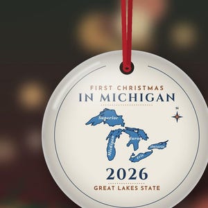 May include: A round white ceramic ornament hanging from a red ribbon. The text reads FIRST CHRISTMAS IN MICHIGAN, 2026, and GREAT LAKES STATE. A blue map of the Great Lakes is centered with a compass rose to the right of the lakes.