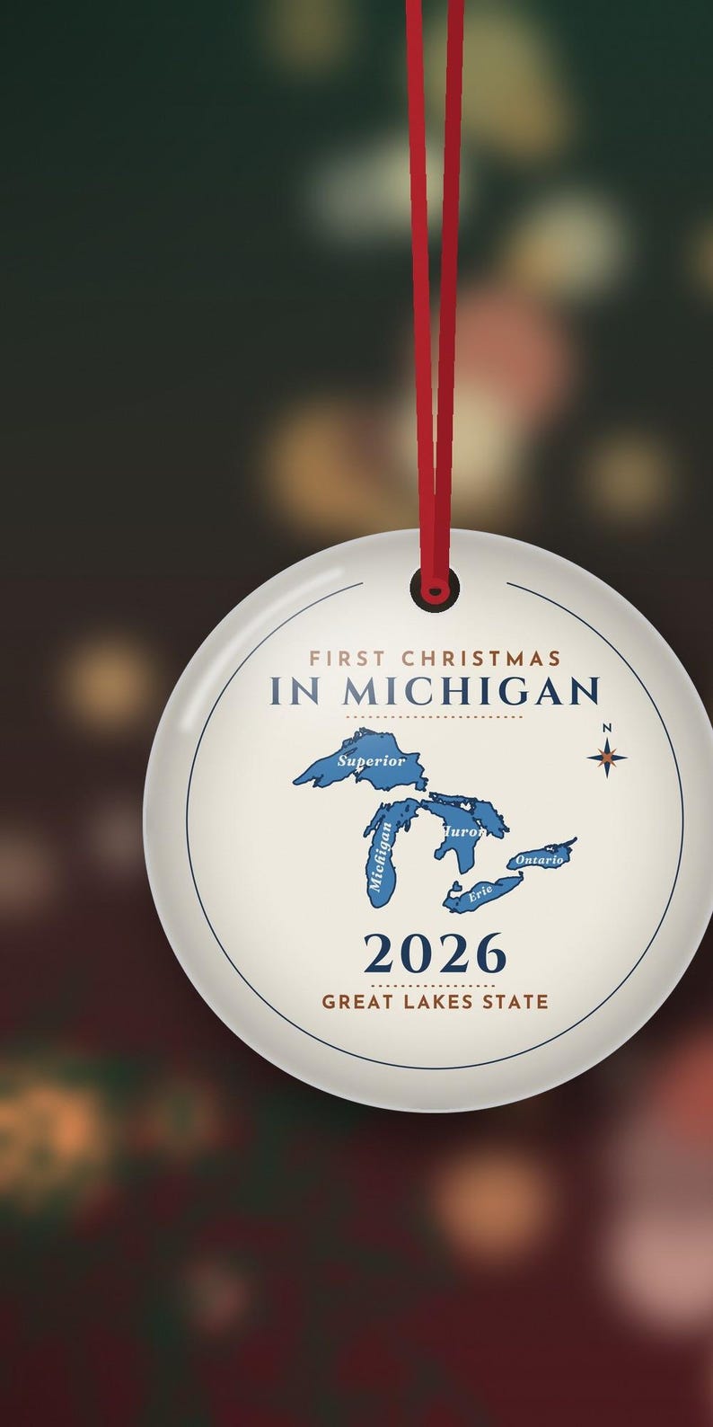 May include: A round white ceramic ornament hanging from a red ribbon. The text reads FIRST CHRISTMAS IN MICHIGAN, 2026, and GREAT LAKES STATE. A blue map of the Great Lakes is centered with a compass rose to the right of the lakes.