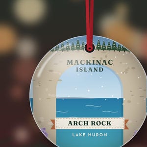 May include: A round, flat ornament hanging from a red ribbon. The design features a graphic illustration of Arch Rock on Mackinac Island, Lake Huron, with a blue sky, water, and a line of green pine trees at the top. The text reads Mackinac Island, Arch Rock, and Lake Huron.