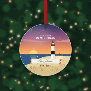 K&ouml;nnte beinhalten: Ein runder, flacher Anh&auml;nger an einem roten Band mit einer Leuchtturm-Illustration bei Sonnenuntergang und dem Text NEW HOME IN MICHIGAN, The Johnsons sowie EST. 2026. Das Design zeigt einen violetten Sternenhimmel, eine Sonne und einen Sandstrand.