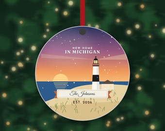 May include: A round, flat ornament hanging from a red ribbon, featuring a lighthouse illustration at sunset with the text NEW HOME IN MICHIGAN, The Johnsons, and EST. 2026. The design includes a starry purple sky, a sun, and a sandy beach.