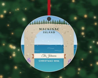 May include: A round ceramic Christmas ornament featuring a graphic illustration of the Arch Rock on Mackinac Island. The design includes a blue sky, a line of green pine trees, blue water, and the text Mackinac Island, The Johnsons, and Christmas 2026.