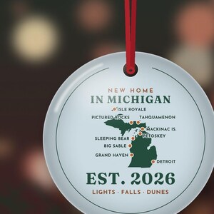 Puede incluir: Un adorno redondo de cer&aacute;mica blanca colgado de una cinta roja. Presenta un mapa verde de Michigan con puntos rojos que marcan lugares como Isle Royale, Pictured Rocks y Detroit. El texto dice: NEW HOME IN MICHIGAN, EST. 2026, LIGHTS &middot; FALLS &middot; DUNES.