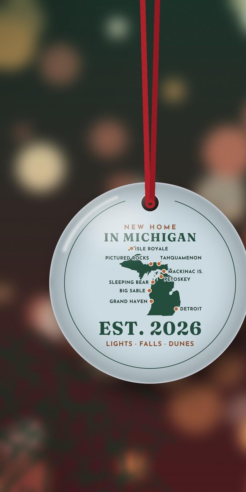 Puede incluir: Un adorno redondo de cer&aacute;mica blanca colgado de una cinta roja. Presenta un mapa verde de Michigan con puntos rojos que marcan lugares como Isle Royale, Pictured Rocks y Detroit. El texto dice: NEW HOME IN MICHIGAN, EST. 2026, LIGHTS &middot; FALLS &middot; DUNES.