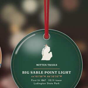Puede incluir: Un adorno redondo de color verde oscuro colgado de una cinta roja. Presenta un mapa blanco de Michigan con una estrella, el texto Mitten Trails, Big Sable Point Light, coordenadas 44&deg;03'28"N 86&deg;30'52"W, First lit 1867, torre de 34 m, Ludington State Park.