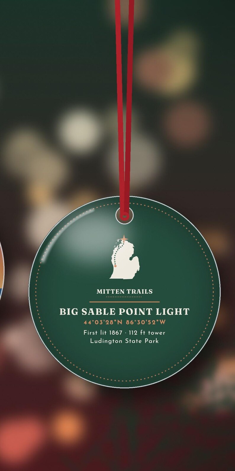 Puede incluir: Un adorno redondo de color verde oscuro colgado de una cinta roja. Presenta un mapa blanco de Michigan con una estrella, el texto Mitten Trails, Big Sable Point Light, coordenadas 44&deg;03'28"N 86&deg;30'52"W, First lit 1867, torre de 34 m, Ludington State Park.