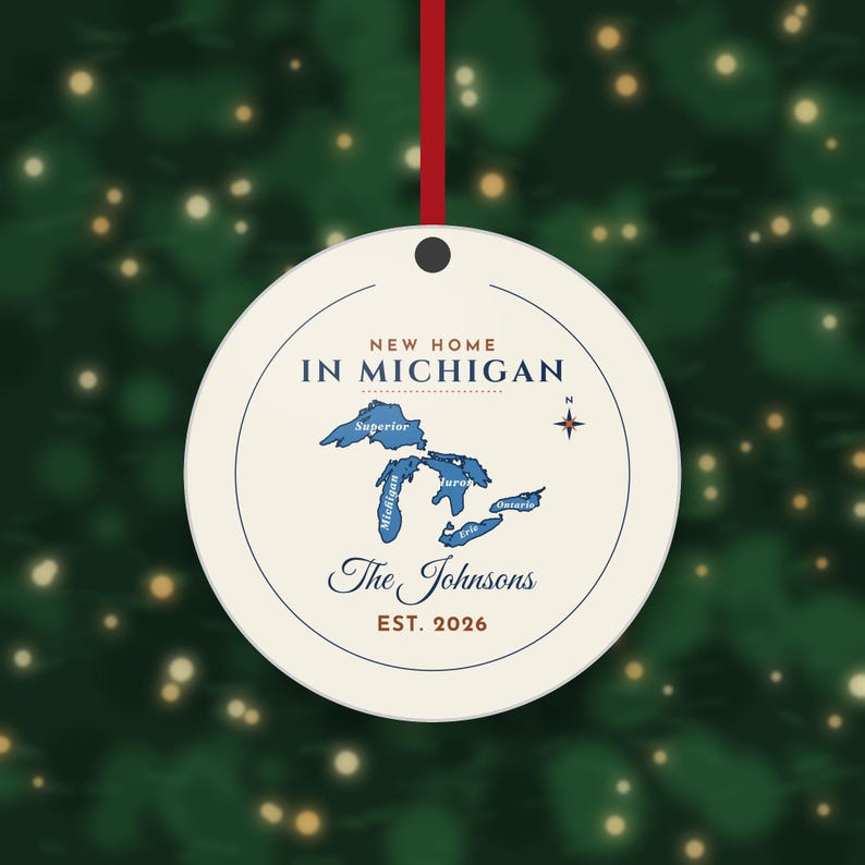 May include: A round, cream-colored ornament hanging from a red ribbon. It features a blue map of the Great Lakes, a compass rose, and the text: NEW HOME IN MICHIGAN, The Johnsons, and EST. 2026. The design is enclosed within a thin circular line.