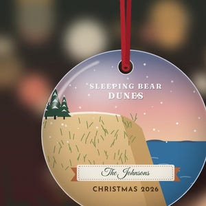 Puede incluir: Un adorno navide&ntilde;o redondo y plano colgado de una cinta roja. El dise&ntilde;o muestra un paisaje de Sleeping Bear Dunes con &aacute;rboles verdes, una colina de arena y agua azul bajo un cielo al atardecer. Texto: SLEEPING BEAR DUNES, The Johnsons, CHRISTMAS 2026.