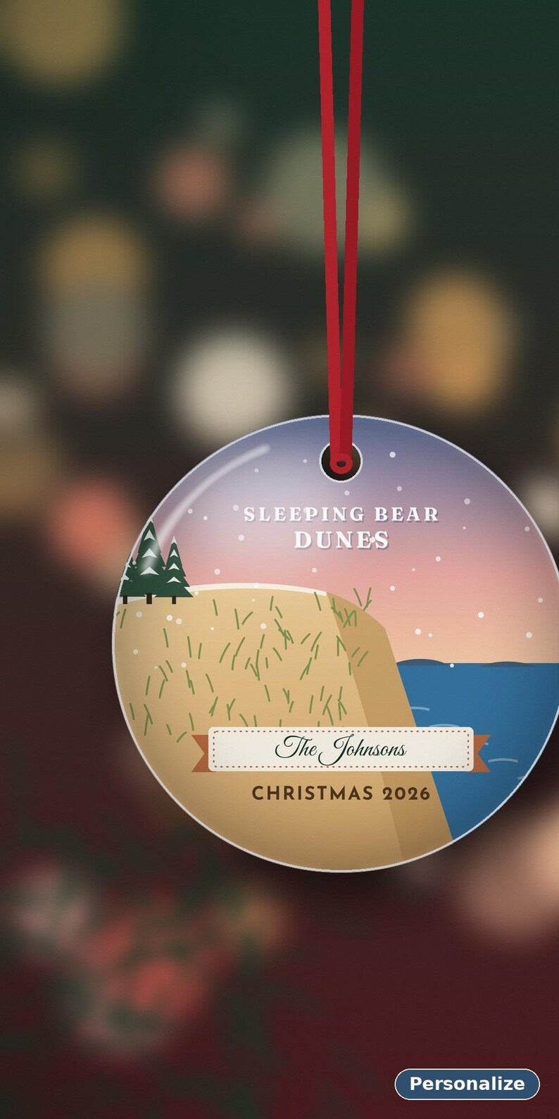 Puede incluir: Un adorno navide&ntilde;o redondo y plano colgado de una cinta roja. El dise&ntilde;o muestra un paisaje de Sleeping Bear Dunes con &aacute;rboles verdes, una colina de arena y agua azul bajo un cielo al atardecer. Texto: SLEEPING BEAR DUNES, The Johnsons, CHRISTMAS 2026.