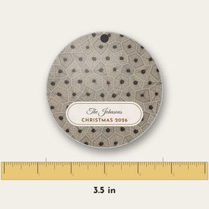 May include: A circular ornament featuring a beige and brown geometric pattern of interconnected polygons with dark centers. A central white horizontal label displays the text The Johnsons CHRISTMAS 2026. A ruler shows a diameter of 3.5 inches.