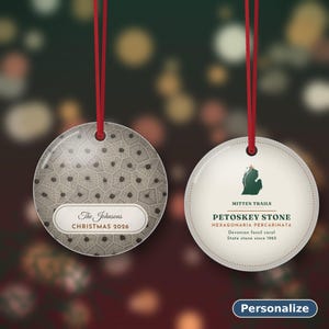 May include: Two round ceramic ornaments with red hanging ribbons. The left ornament features a gray hexagonal pattern and the text The Johnsons CHRISTMAS 2026. The right ornament displays a green Michigan map, MITTEN TRAILS, PETOSKEY STONE, HEXAGONARIA PERCARINATA, Devonian Fossil coral, and State stone since 1965.