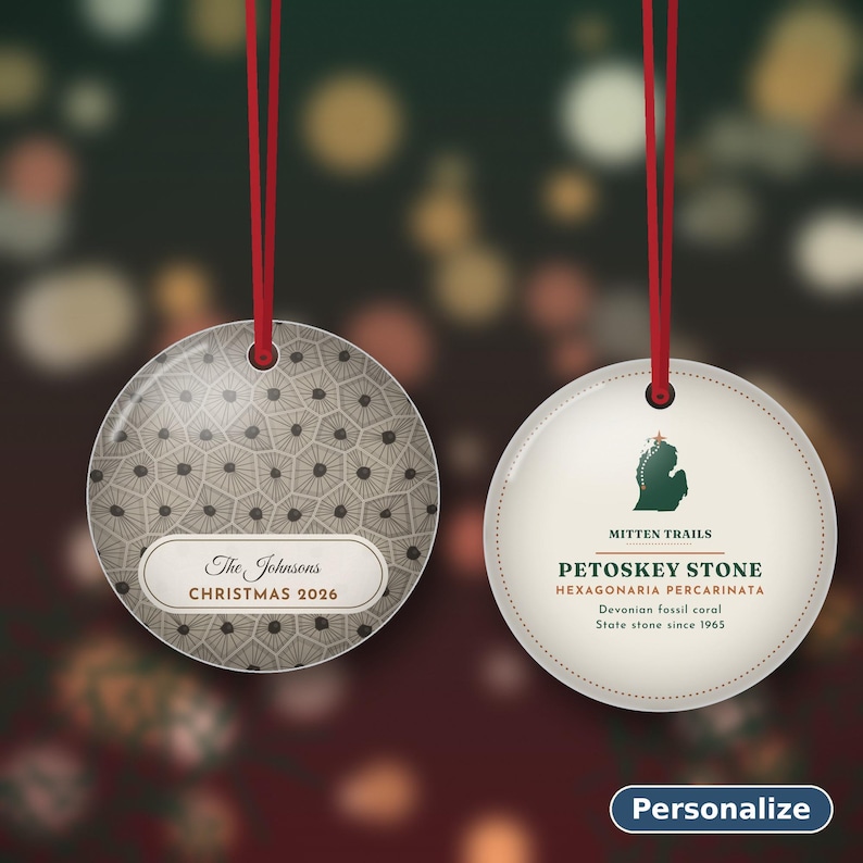 May include: Two round ceramic ornaments with red hanging ribbons. The left ornament features a gray hexagonal pattern and the text The Johnsons CHRISTMAS 2026. The right ornament displays a green Michigan map, MITTEN TRAILS, PETOSKEY STONE, HEXAGONARIA PERCARINATA, Devonian Fossil coral, and State stone since 1965.