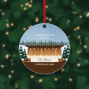 May include: A round Christmas ornament featuring a graphic illustration of Tahquamenon Falls with evergreen trees, a light blue sky, and white snow. The text reads Tahquamenon Falls, The Johnsons, and Christmas 2026, hanging by a red ribbon.