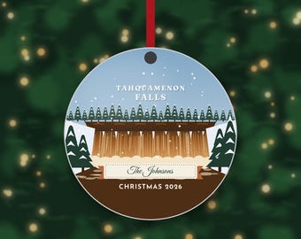 May include: A round Christmas ornament featuring a graphic illustration of Tahquamenon Falls with evergreen trees, a light blue sky, and white snow. The text reads Tahquamenon Falls, The Johnsons, and Christmas 2026, hanging by a red ribbon.