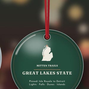 May include: A round, dark green ornament hanging from a red ribbon. It features a white silhouette of Michigan with a star, the text "MITTEN TRAILS", "GREAT LAKES STATE", and "Pinned: Isle Royale to Detroit Lights &middot; Falls &middot; Dunes &middot; Islands".