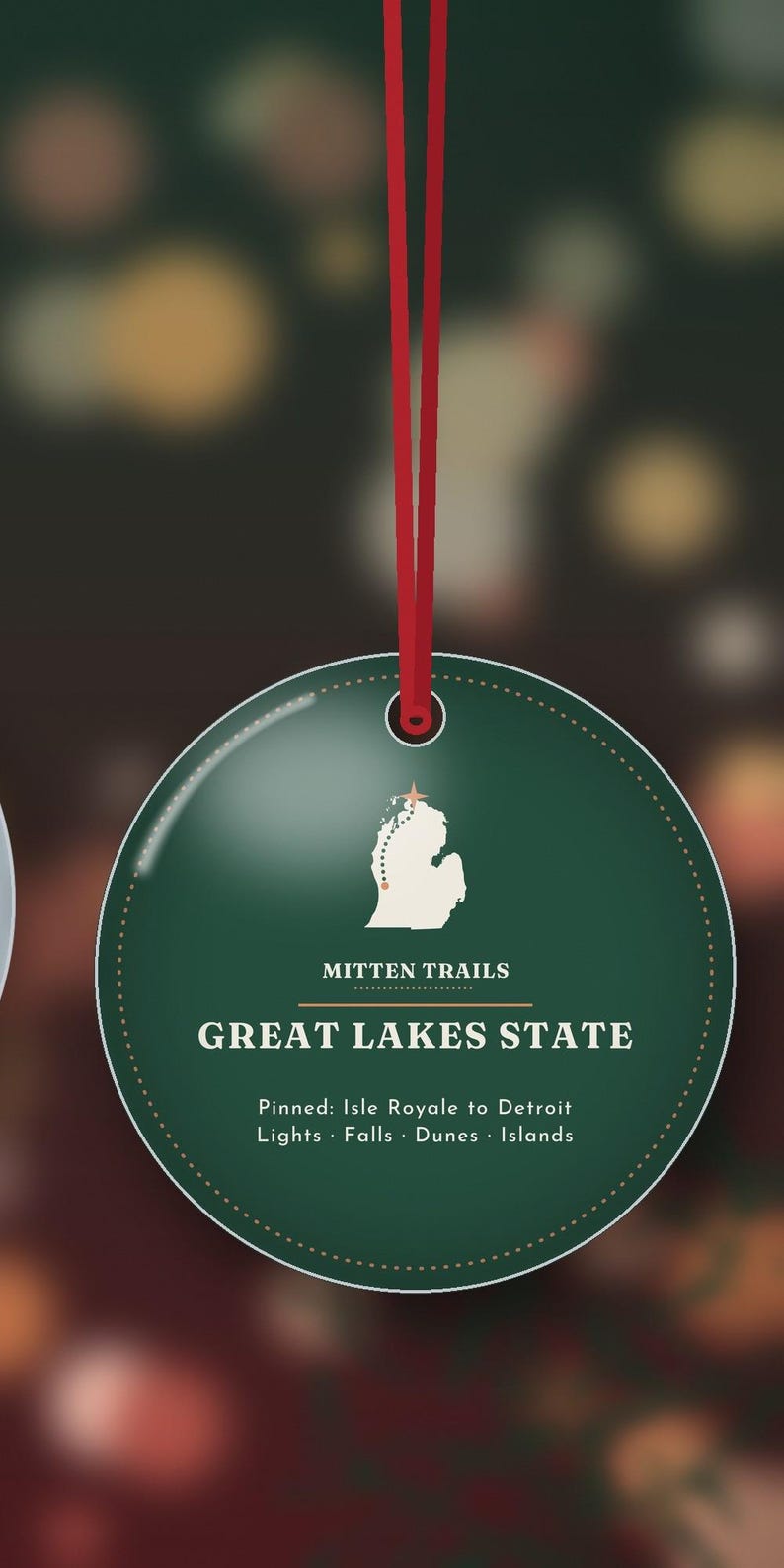 May include: A round, dark green ornament hanging from a red ribbon. It features a white silhouette of Michigan with a star, the text "MITTEN TRAILS", "GREAT LAKES STATE", and "Pinned: Isle Royale to Detroit Lights &middot; Falls &middot; Dunes &middot; Islands".