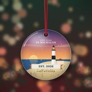 Puede incluir: Un adorno redondo de cerámica colgado de una cinta roja, con una escena de atardecer con un faro, el texto NEW HOME IN MICHIGAN, EST. 2026 y LAKE MICHIGAN. El diseño incluye una playa de arena, agua azul y un cielo de color púrpura.